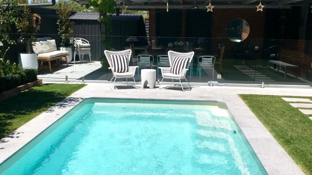 Modern backyard swimming pool with glass fence, white lounge chairs, and green garden